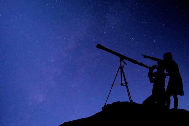 Two people looking through telescopes beneath a field of stars